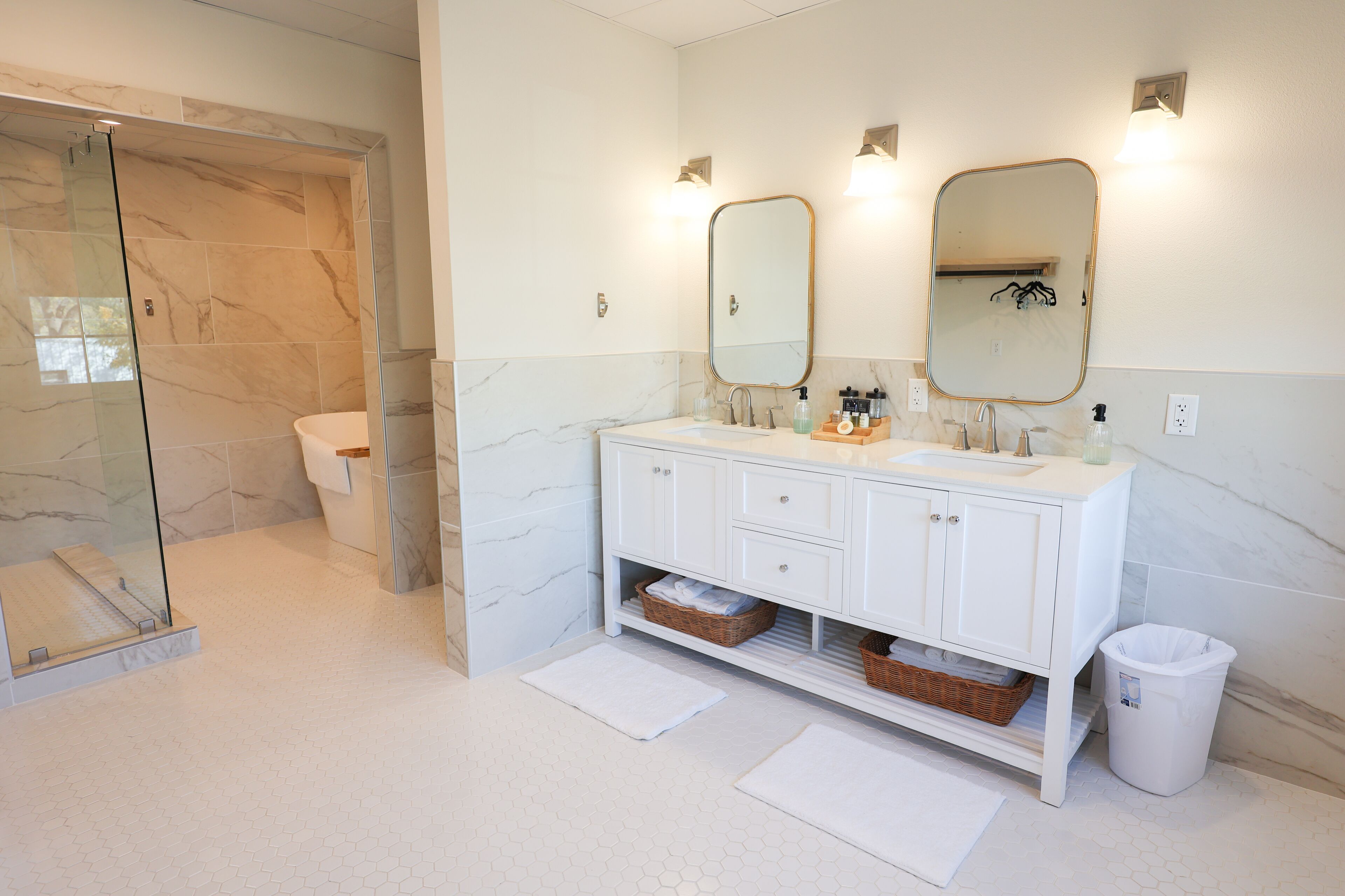 A bathroom with a glass shower enclosure, a white sink with a marble countertop, two wall-mounted mirrors, and a white trash bin.