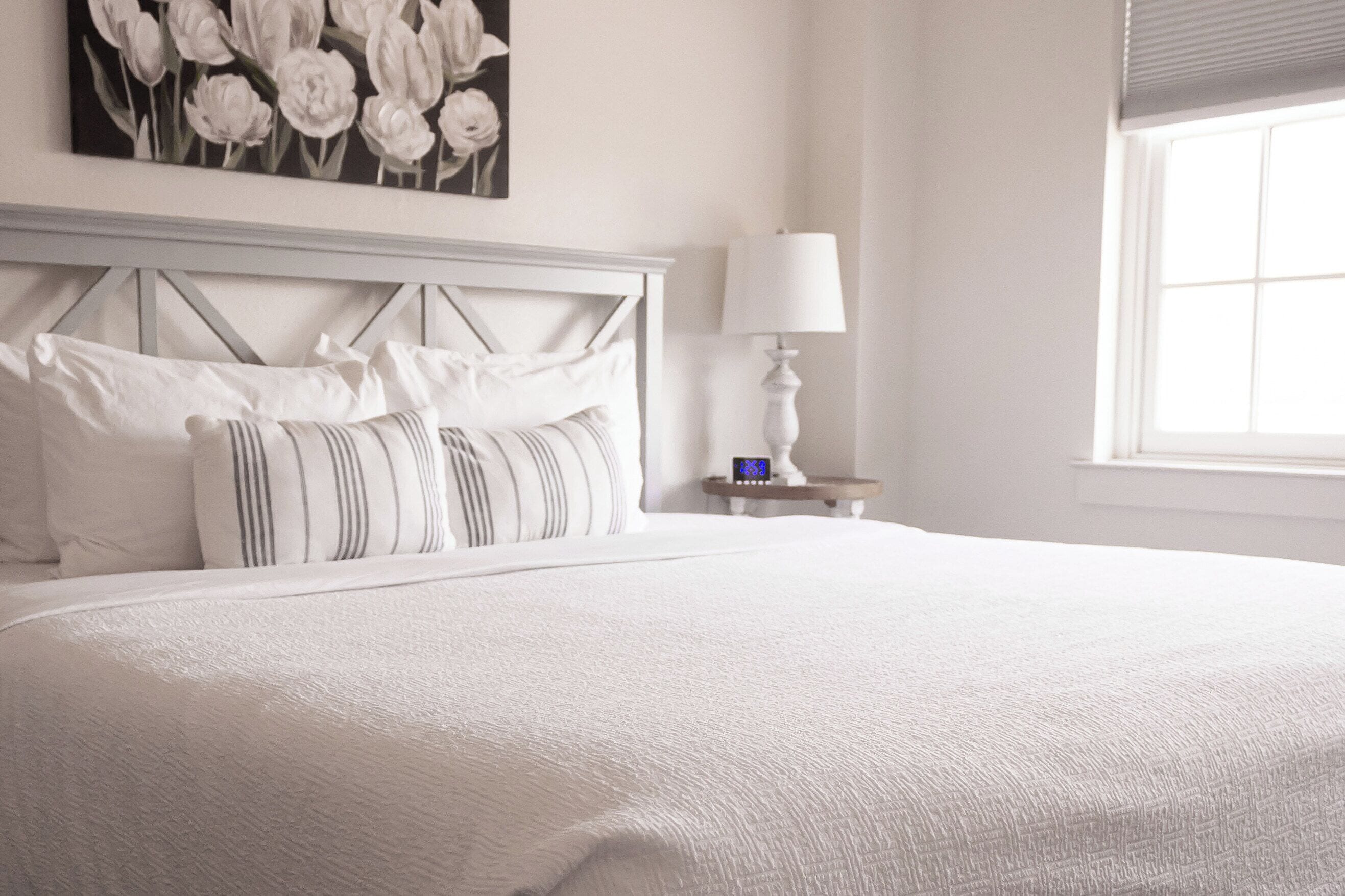 A neatly made bed with white bedding and striped pillows, a bedside table with a lamp, and a wall-mounted painting of flowers.