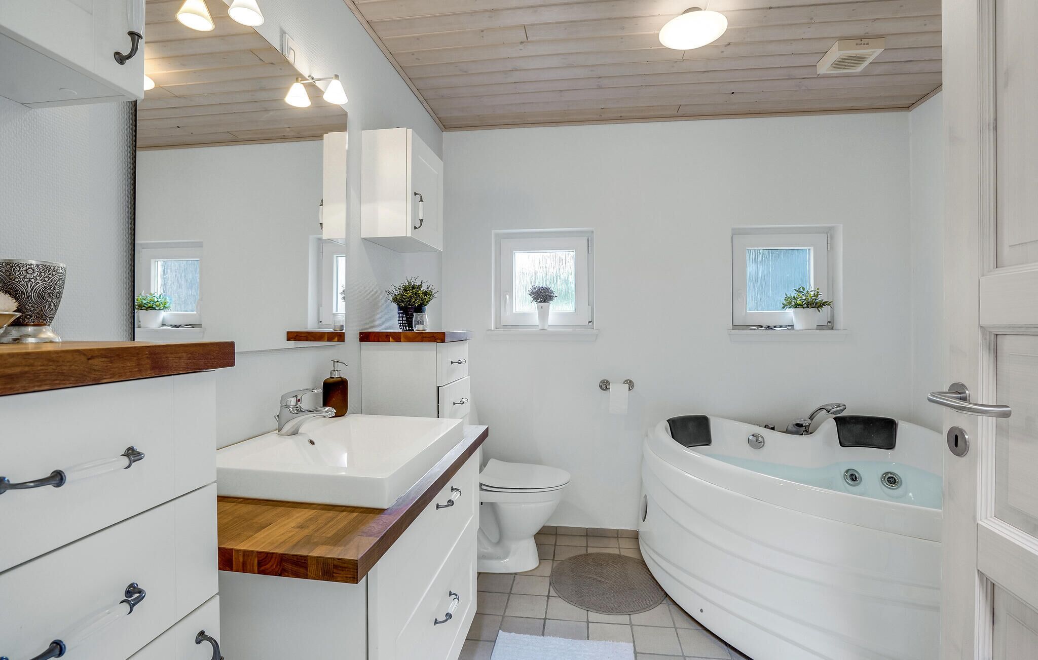 Une salle de bain moderne avec un lavabo, des toilettes et une baignoire, tous de couleur blanche. On y trouve deux fenêtres ornées de plantes, des meubles en bois et un miroir.