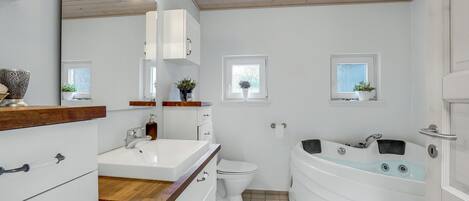 A modern bathroom with a white sink, toilet, and bathtub. There are two windows with plants, wooden cabinets, and a mirror.