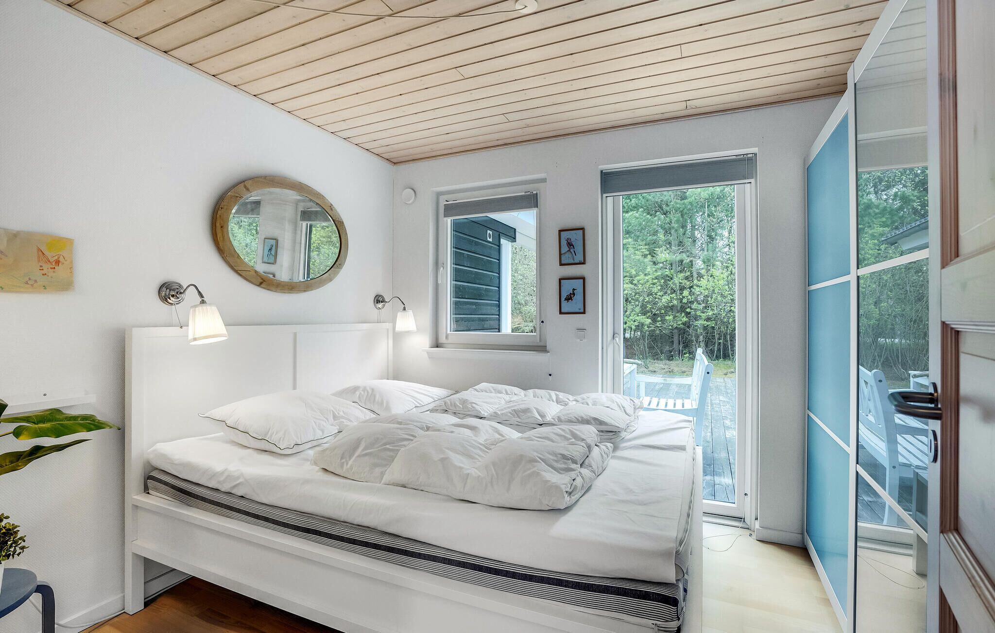 A bedroom with a bed, a round mirror, two wall-mounted lamps, and a window with a view of trees.