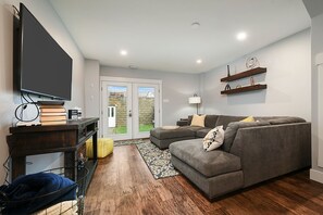 Un salon avec un canapé gris, un téléviseur à écran plat fixé sur un meuble en bois, et une bibliothèque contenant des livres et des objets décoratifs.