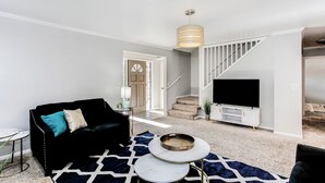 A living room with a black sofa, a round coffee table, and a TV on a white stand. There is a staircase leading to another level. - Spacious 2,500 Sq Ft Wichita Home -3 Showers-2 Tubs-Near Base & Arena (Wichita)