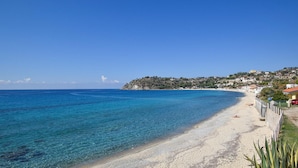 Une plage côtière aux eaux bleues et limpides, bordée d’un rivage sablonneux et adossée à une colline où se trouvent des bâtiments.