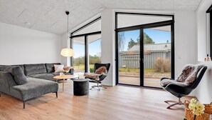 Un salon moderne avec un canapé d’angle gris, une table basse ronde et deux fauteuils. De grandes portes vitrées donnent sur un jardin.