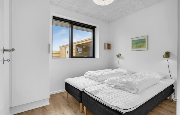 A bedroom with two beds, a window with a view of a building, a framed picture on the wall, and a ceiling light fixture.
