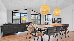 Un vaste espace de vie comprenant une grande table à manger en bois entourée de chaises, un canapé gris et une grande fenêtre donnant sur l’extérieur.