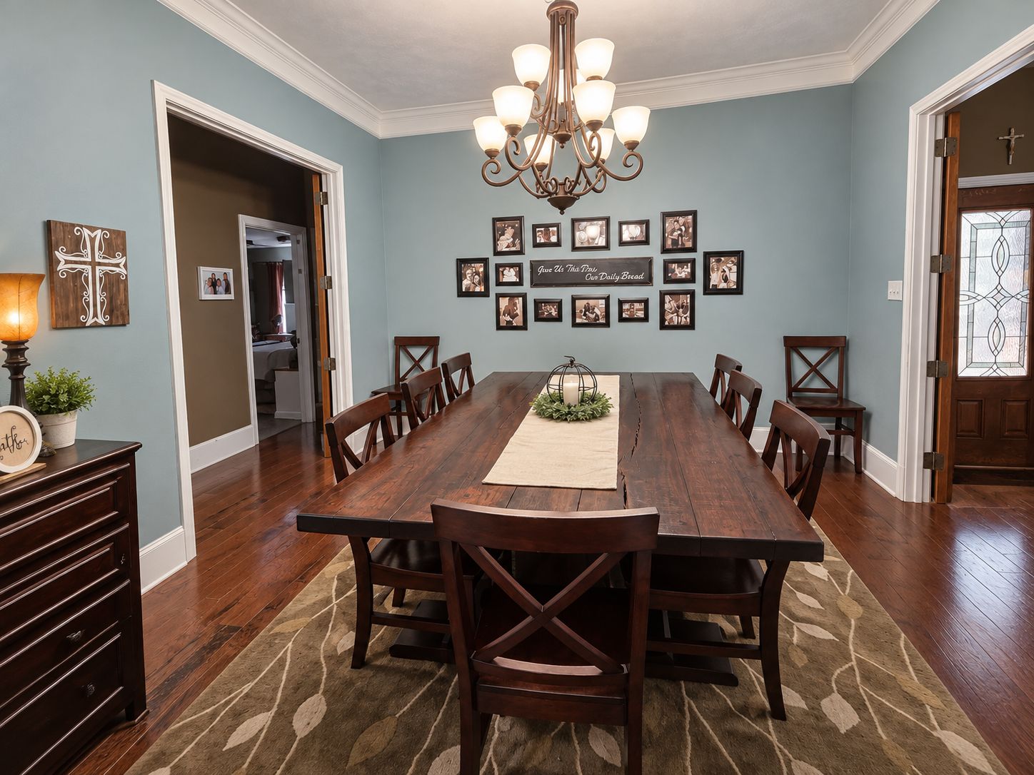 A dining room with a long wooden table, surrounded by chairs, a chandelier, framed photos on the wall, and a cross decoration.