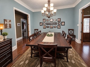 Uma sala de jantar com uma mesa de madeira comprida, rodeada por cadeiras, um candelabro, fotografias emolduradas na parede e uma cruz decorativa.