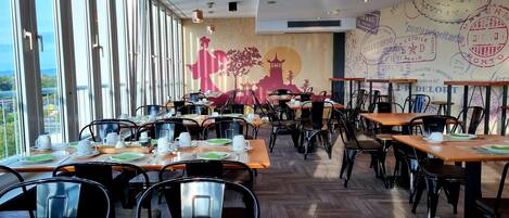 A restaurant with multiple tables and chairs, large windows, and a mural on the wall.