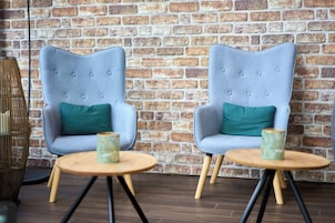 Dos sillones tapizados en gris con cojines verdes, una mesa redonda de madera con un jarrón verde y una pared de ladrillo de fondo.