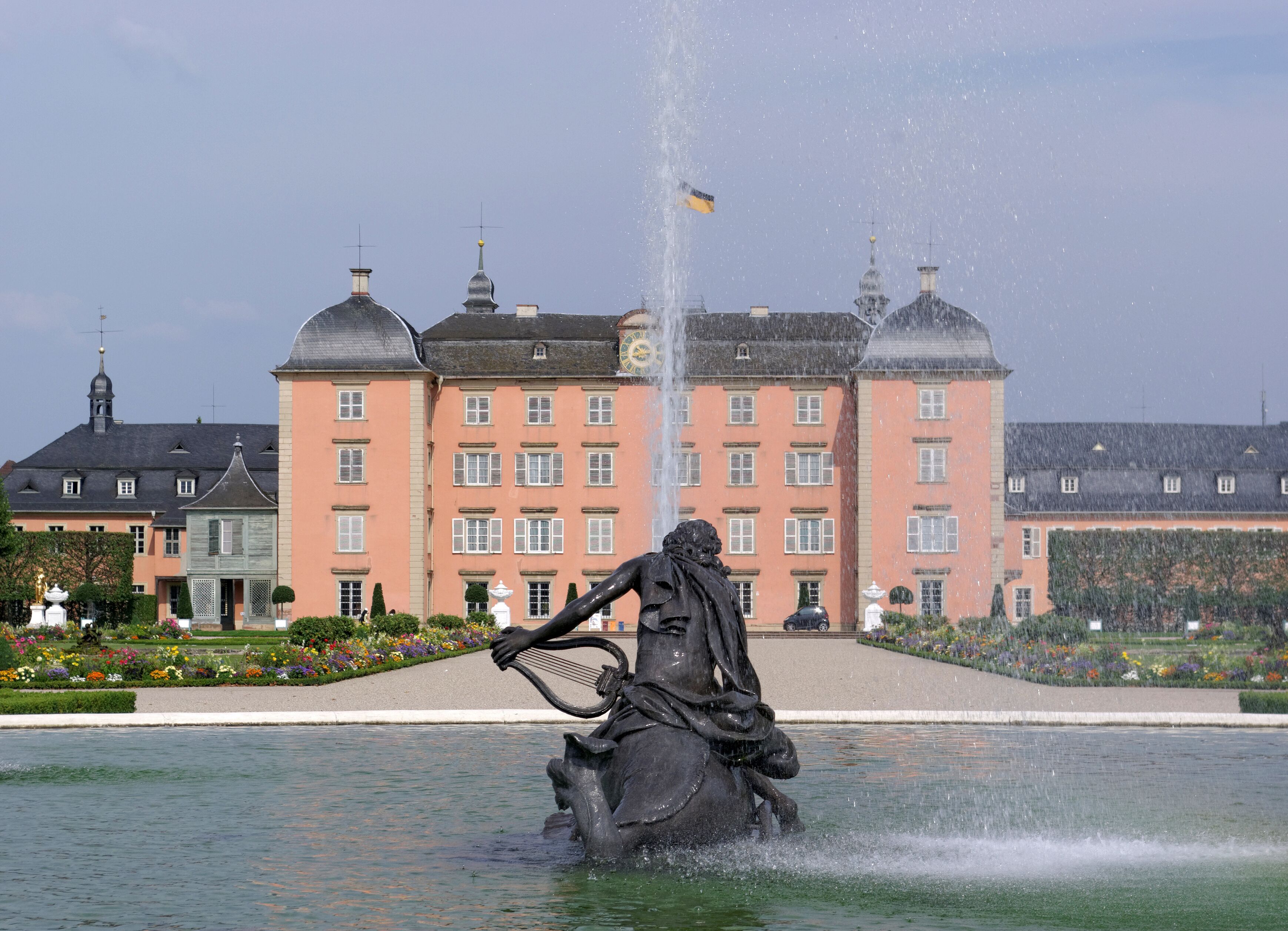 A fountain with a statue of a man playing a lyre, surrounded by water. In the background, there is a large pink building with multiple windows and domes.