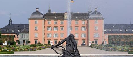 A fountain with a statue of a man playing a lyre, surrounded by water. In the background, there is a large pink building with multiple windows and domes.