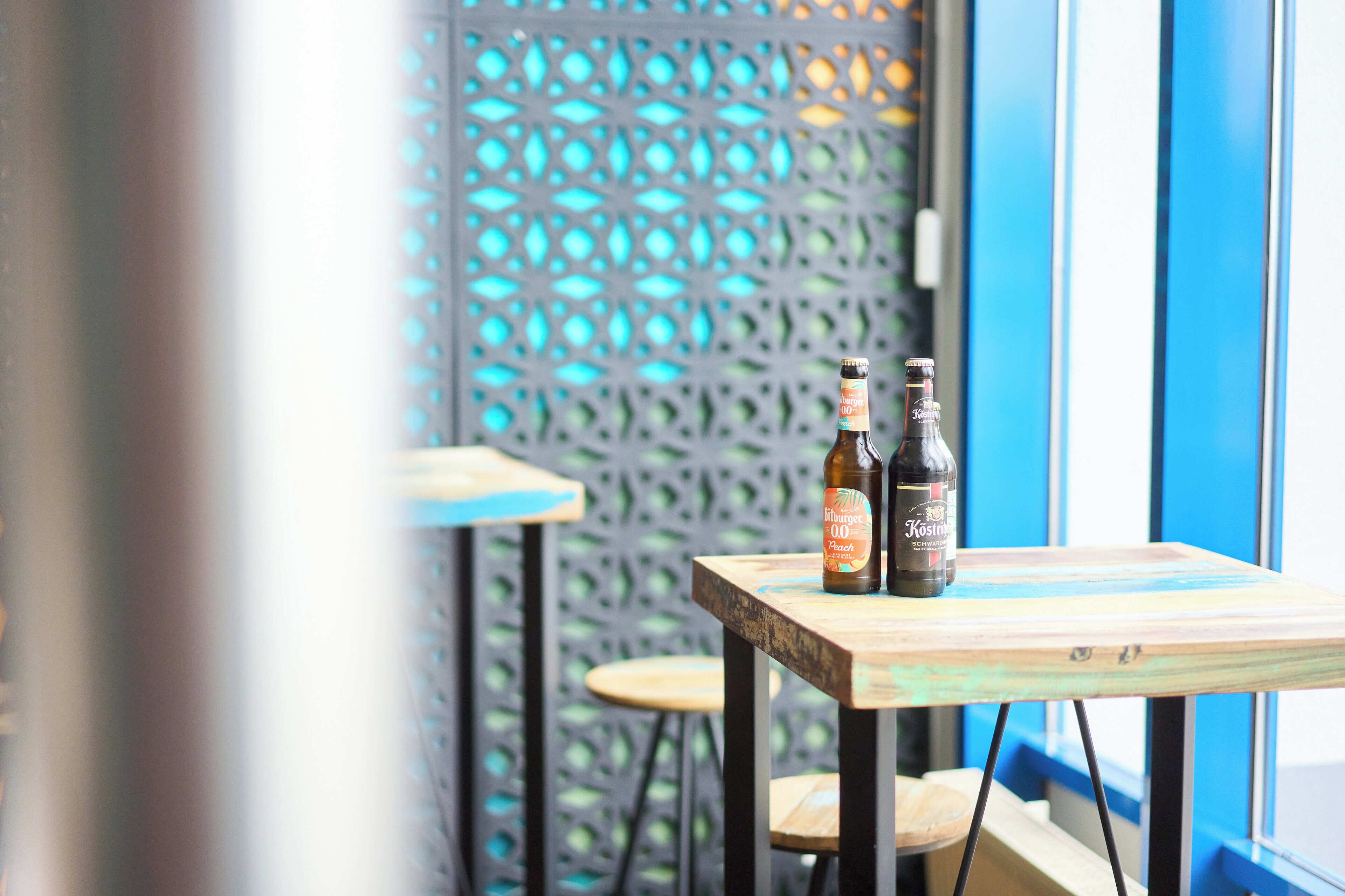 A table with two bottles of beer and a wooden chair in a room with a blue and white patterned wall.