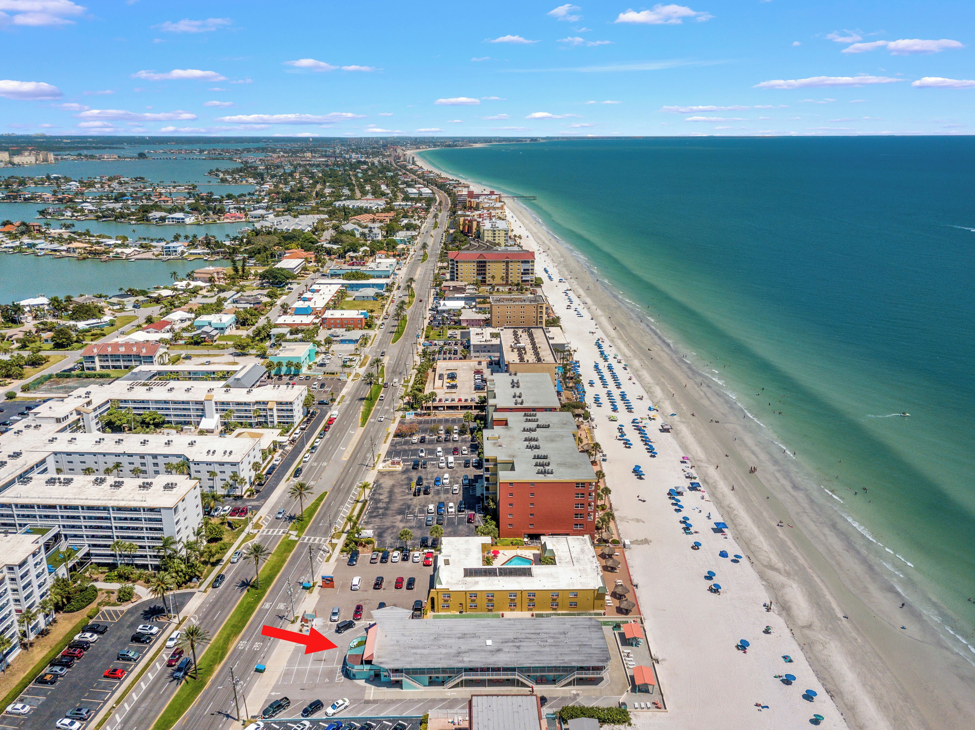 Vista aérea de una ciudad costera con una extensa playa, un puerto deportivo y una mezcla de edificios comerciales y residenciales.