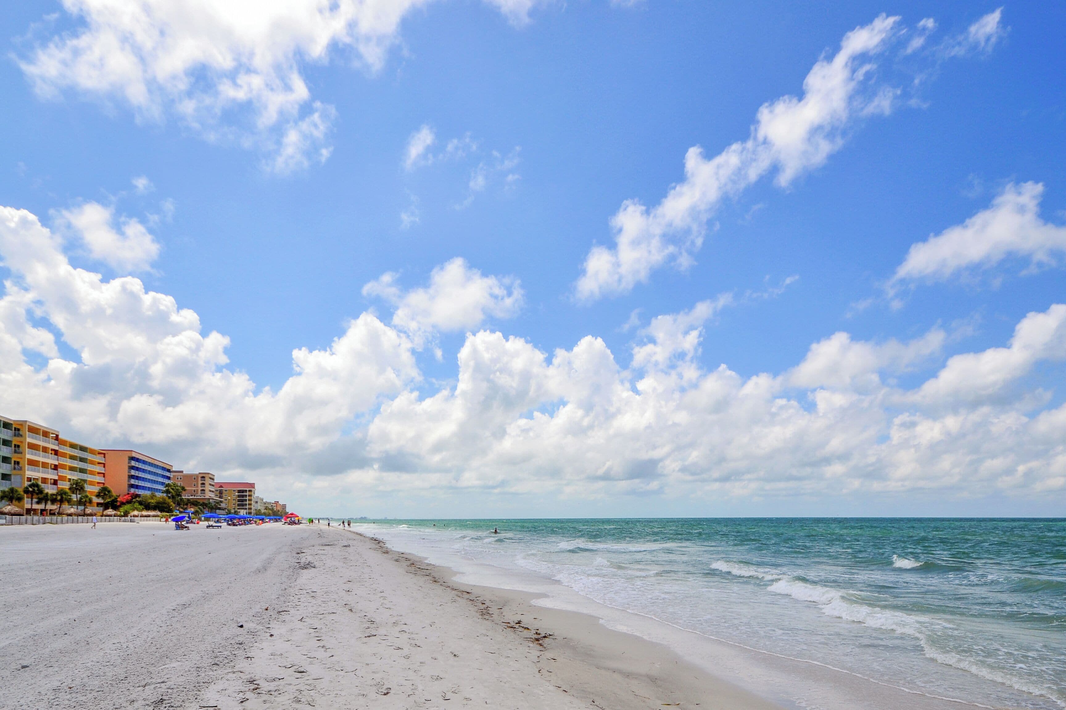 A beach with a clear sky, sandy shore, and buildings in the background.