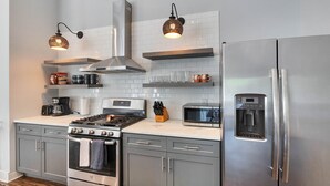 A modern kitchen with stainless steel appliances, including a stove, oven, microwave, and refrigerator. There are two hanging pendant lights, a coffee maker, and various kitchen items on the countertops.
