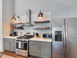 Uma cozinha moderna com eletrodomésticos de aço inoxidável, incluindo fogão, forno, micro-ondas e geladeira. Há duas luminárias pendentes, uma cafeteira e diversos utensílios de cozinha nas bancadas.