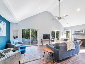 Uma sala de estar ampla com um sofá cinzento grande, poltronas azuis e uma mesa de centro de madeira. Há uma televisão e uma lareira.