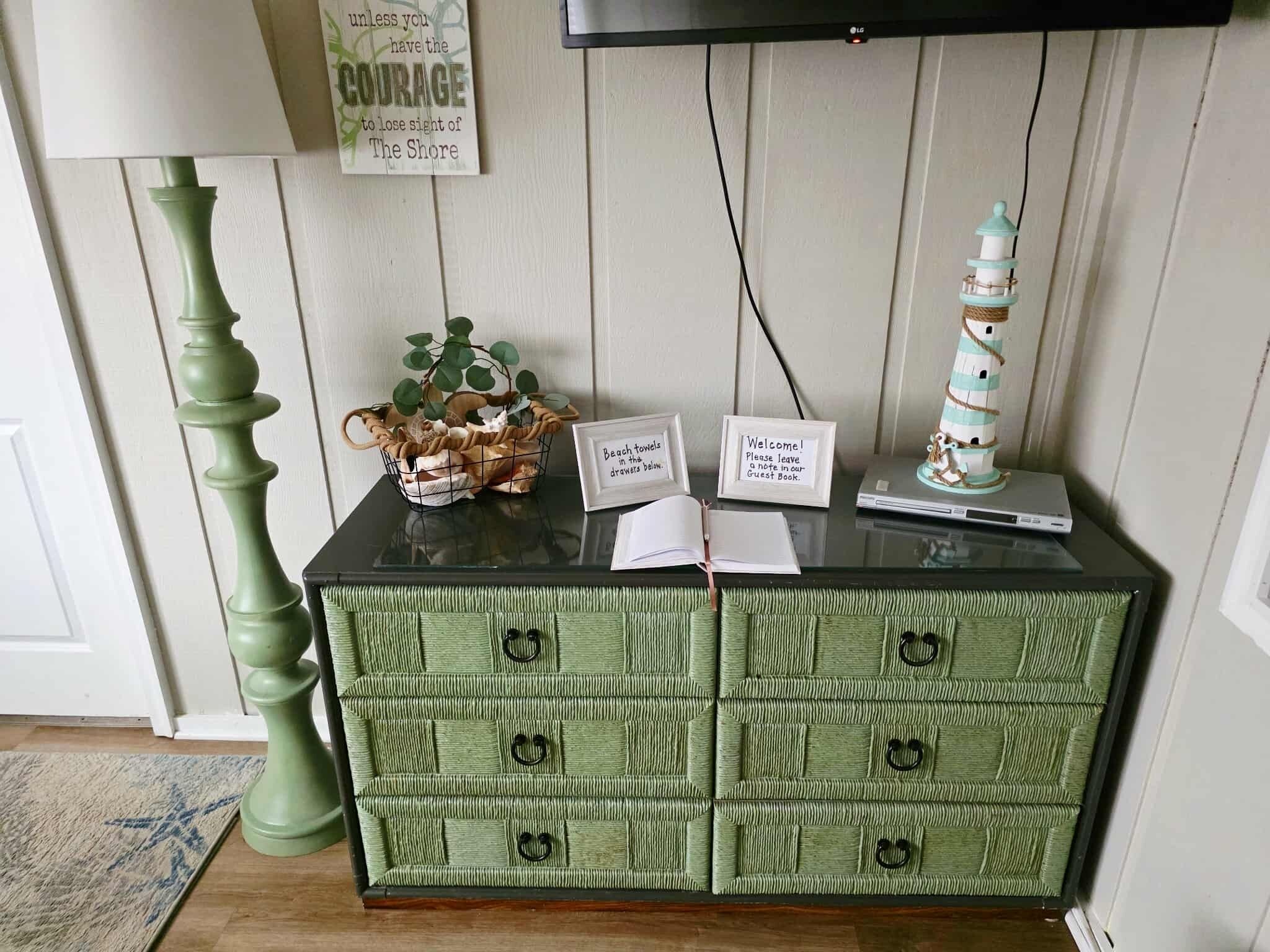 A green chest of drawers with a lamp, a lighthouse decoration, and a sign with a motivational quote.