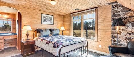 A cozy bedroom with a wooden ceiling, a large bed with a quilt, two bedside lamps, a stone wall, and a view of the outdoors.