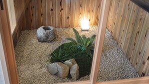 Ein kleiner Innenbereich mit Garten, der eine Holzfassade, eine Lichtquelle sowie verschiedene Pflanzen und Steine zeigt.