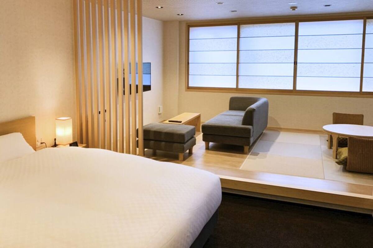 A hotel room with a large bed, a desk, a chair, a small table, and a window with blinds.