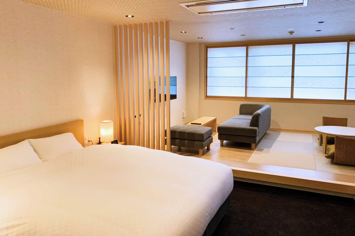 A hotel room with a large bed, a seating area with a table, and a window with blinds.