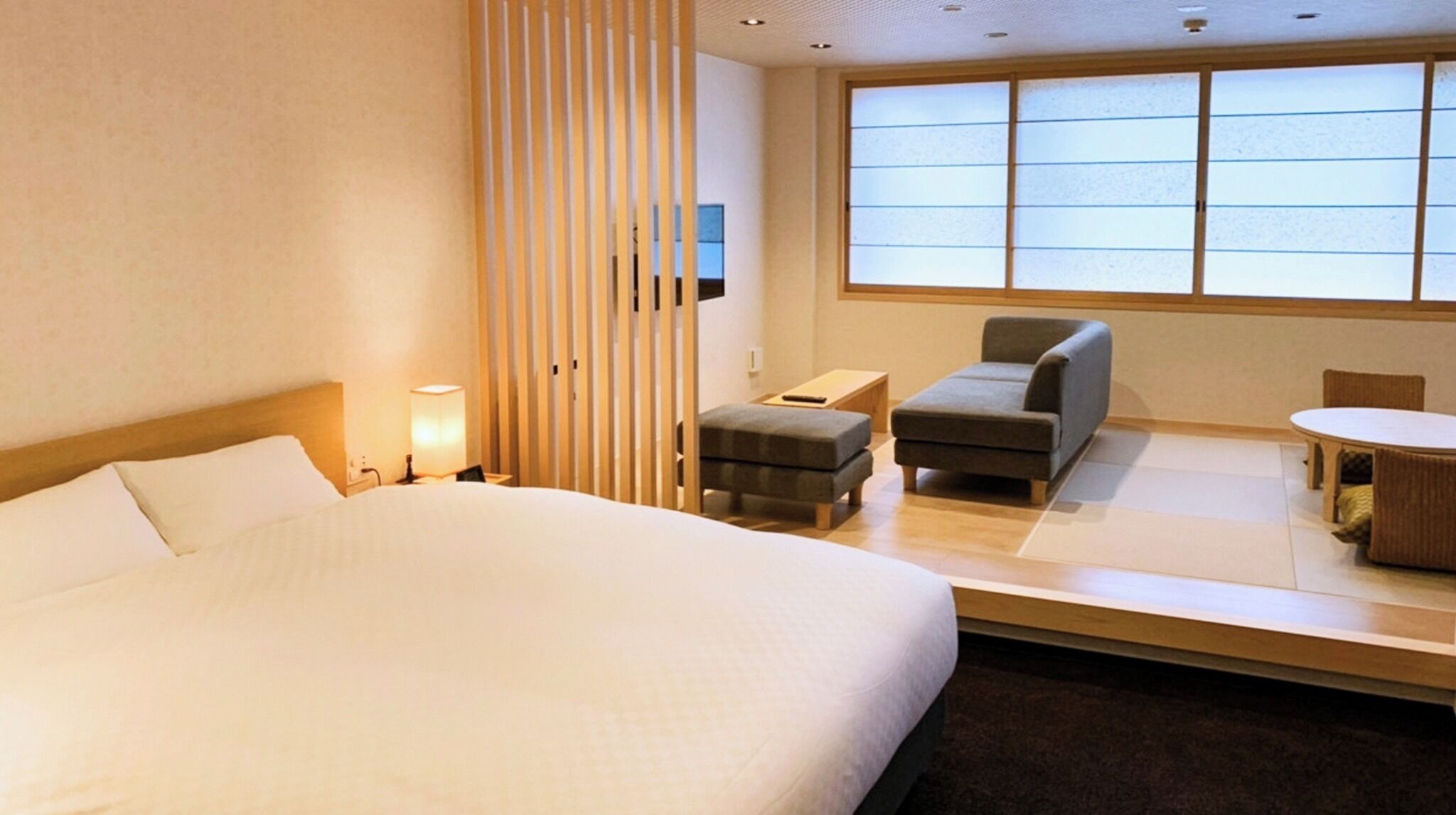 A hotel room with a large bed, a small sofa, a table, and a window with blinds.
