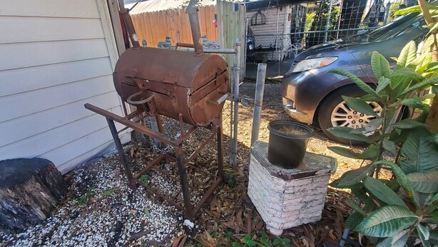 Un entorno rústico al aire libre con un barril metálico, un horno de ladrillo y un coche estacionado cerca.