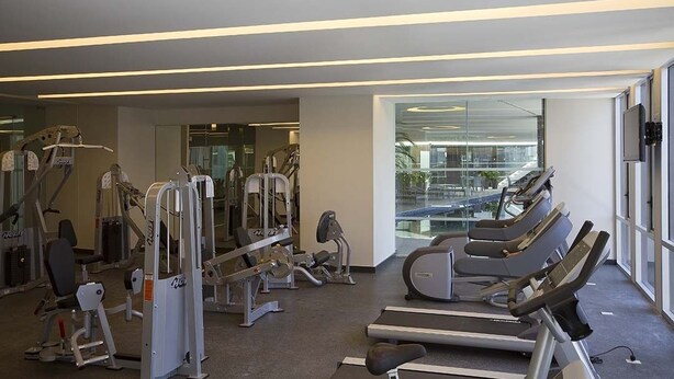 Une salle de sport équipée de tapis de course, d’appareils de musculation et dotée d’un mur en verre donnant sur la ville.