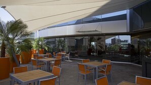 Un café en plein air avec des tables et des chaises, de grands pots de plantes et un bâtiment moderne en arrière-plan.