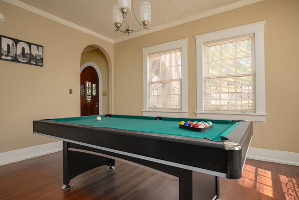 Une table de billard dans une pièce dotée d’un plancher en bois, d’un lustre et de deux fenêtres équipées de stores.