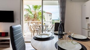 Zona pranzo con tavolo apparecchiato per due, televisione a parete e vista su palme e edifici attraverso una porta scorrevole in vetro.