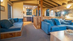 Ein gemütliches Blockhaus-Interieur mit Holzbalkendecke, einer Couch, einem Couchtisch und einem durch einen offenen Durchgang einsehbarer Küchenbereich.