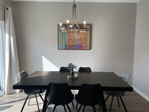 A modern dining area with a large rectangular table, six chairs, a chandelier, and a colorful abstract painting on the wall.