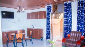 Ein modernes Wohnzimmer mit Glastisch, Ledersofa und einem Essbereich mit Holzkästen sowie einer kleinen Küche im Hintergrund.