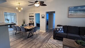 A modern living room with a dark brown sofa, a dining area with a wooden table and grey chairs, a ceiling fan, and a painting of a beach scene. - New Listing!! Gulf Coast beaches, 14 min to ManasotaKey, private heated pool! (Englewood)