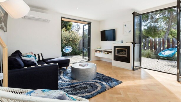 Un soggiorno moderno con un divano nero, un poggiapiedi rotondo, un tappeto blu, una poltrona in rattan bianco, una TV montata a parete e un caminetto.