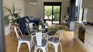 Un soggiorno moderno con tavolo da pranzo in vetro, sedie bianche, un divano blu e una grande finestra vista su vegetazione.