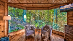 Acogedora habitación tipo cabaña con paredes y suelos de madera, dos sillones, una mesita y un ventanal con vistas al bosque.