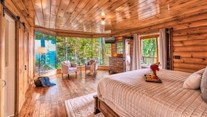 Un dormitorio de cabaña de madera con una cama grande, un aparador, una silla y vistas al bosque.