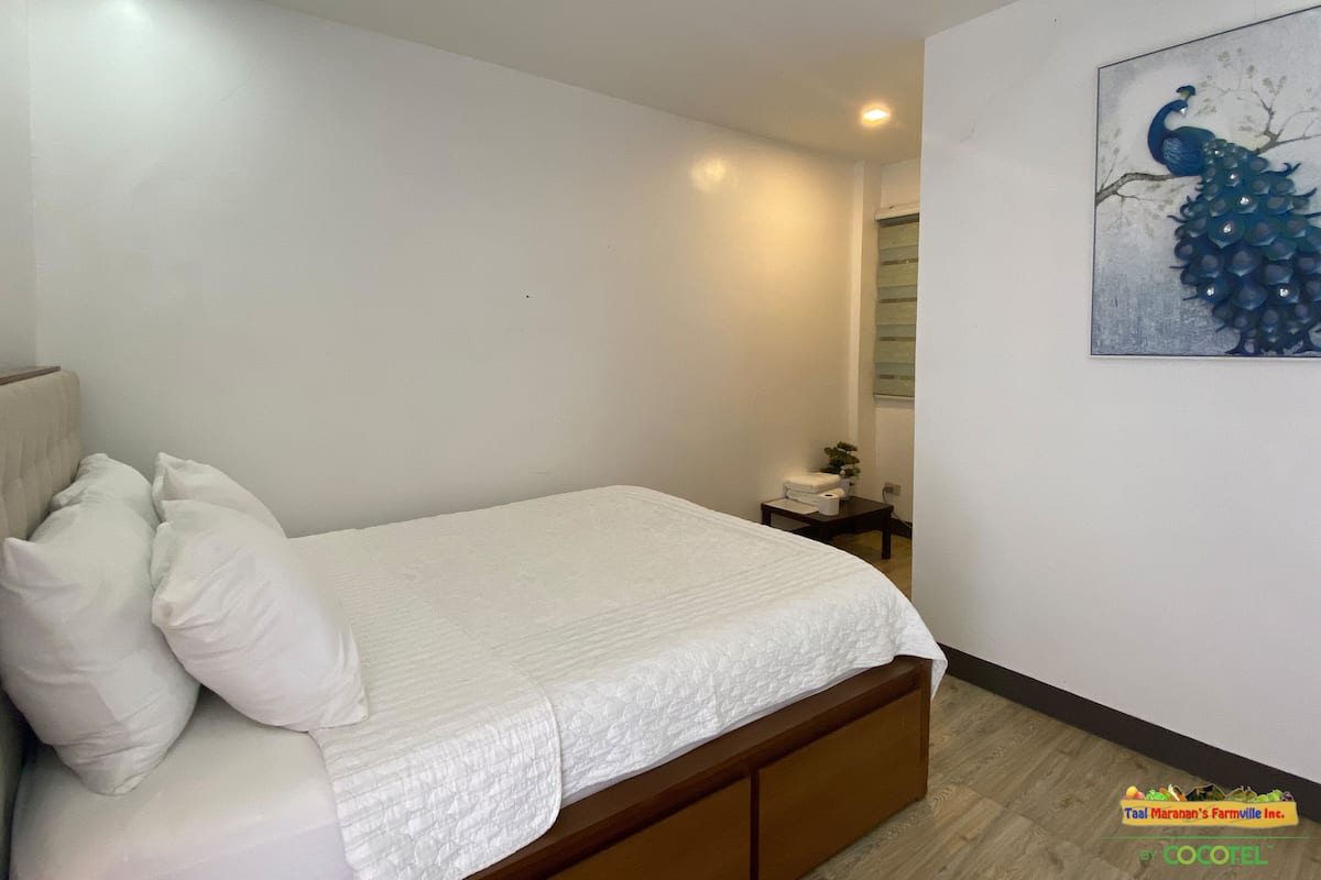 A bedroom with a bed, pillows, a nightstand, a painting, and a window with blinds.