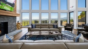 Ein großzügiges Wohnzimmer mit einer großen Couch, einem Couchtisch und einem Kamin. Mehrere Fenster bieten Blick auf die Berge.