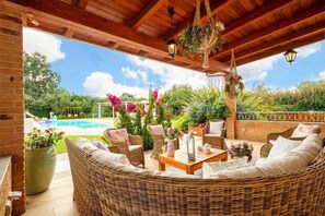 A covered outdoor seating area with wicker furniture, a pool, and lush greenery in the background.