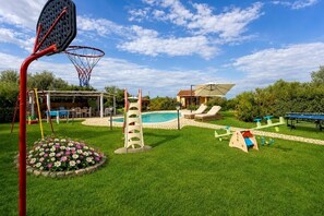 A backyard with a basketball hoop, a swimming pool, lounge chairs, and a climbing structure.
