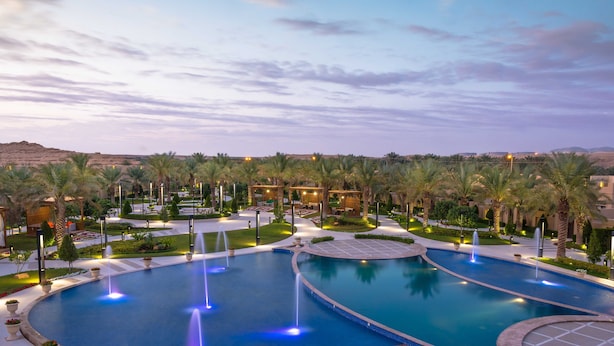 A large outdoor swimming pool with multiple fountains, surrounded by palm trees and well-lit pathways. - Dorat Najd Resort (Diryah)
