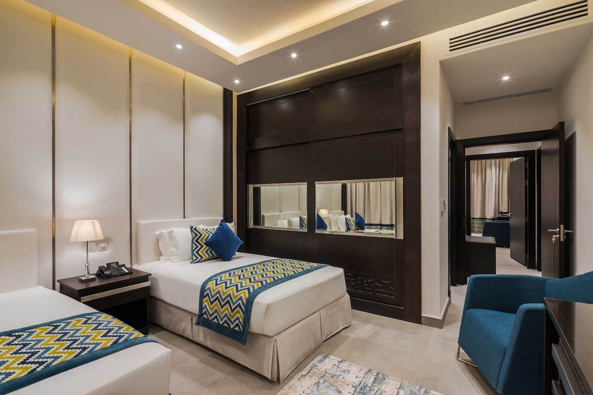 Une chambre d’hôtel moderne avec un grand lit, une table de chevet avec une lampe, un miroir et un fauteuil bleu.