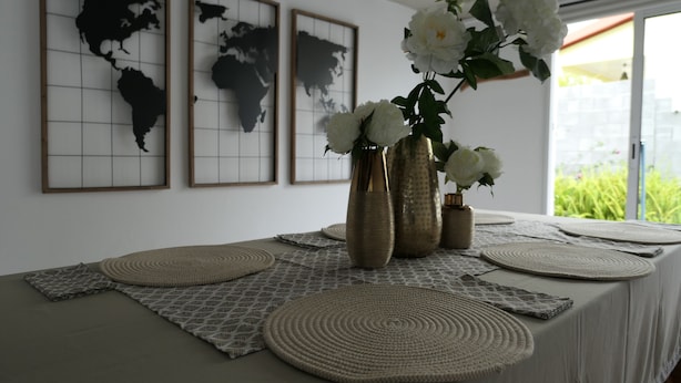 Ein Tisch mit gewebten Tischsets, drei Vasen mit weißen Blumen und gerahmte Weltkarten an der Wand.