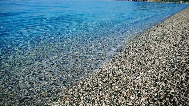 Ein Kiesstrand mit klarem, blauem Wasser und einem Blick auf eine Küstenstadt in der Ferne.
